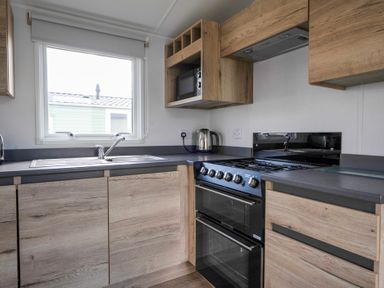 A kitchen with wooden cabinets a window a sink a microwave a kettle and a black oven at Kingfisher Caravan - Billing Aquadrome in Billing Aquadrome Holiday Park