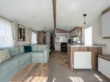 A living area with a light green sofa and wooden coffee table next to a kitchen with wooden cabinets and appliances at Stillwater Retreat - Billing Aquadrome Holiday Park