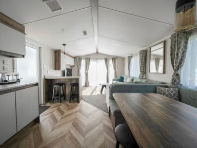 An open plan living space with a kitchen area including stools and a toaster a dining area with a wooden table and cushioned seating and a seating area with sofas and curtains at Stillwater Retreat Billing Aquadrome Holiday Park