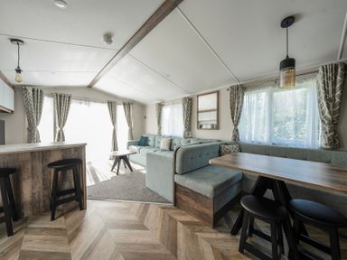 A living room with a dining area a sofa a coffee table bar stools and windows with curtains at Stillwater Retreat - Billing Aquadrome in Billing Aquadrome Holiday Park