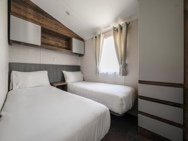 A bedroom with two single beds a window with curtains and built in storage cabinets at Stillwater Retreat in Billing Aquadrome Holiday Park