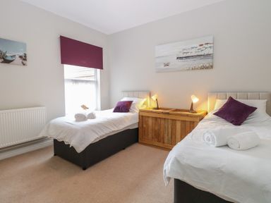 A bedroom with two single beds with white bedding and purple pillows a wooden chest with two lamps and wall art above at 34 North Denes Road Great Yarmouth