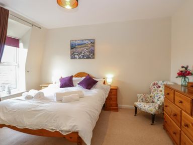 A bedroom with a wooden bed with white bedding and purple pillows wooden nightstand and dresser a floral armchair and a window at 34 North Denes Road in Great Yarmouth