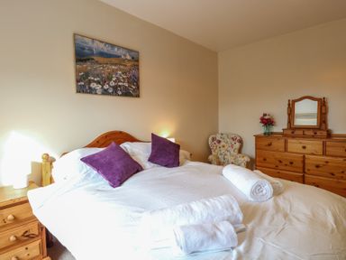 A bedroom with a bed with white sheets and purple pillows a wooden dresser with a mirror and a floral armchair at 34 North Denes Road in Great Yarmouth