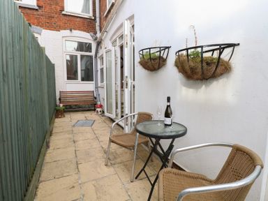 An outdoor patio area with a small round table with a bottle and two glasses and two chairs next to a white wall with two hanging planters and a wooden bench near a brick building at 34 North Denes Road Great Yarmouth