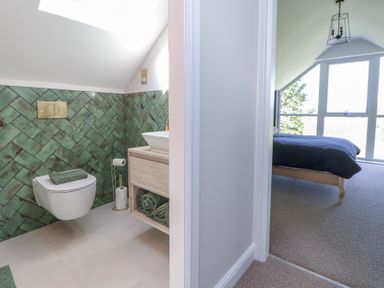 A bathroom with green tiles and a toilet next to a bedroom with a bed near large windows at Ty Blodau near Prestatyn