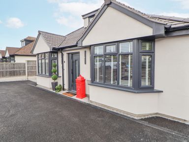 Exterior view of a single-story house with grey window frames a red post box near the black front door and potted plants at Ty Blodau near Prestatyn