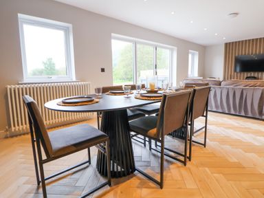 A dining table with six chairs and place settings in a room with windows and a sofa at Ty Blodau near Prestatyn