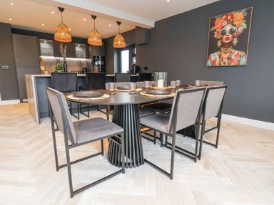 A dining room with a round table and six chairs and a kitchen area with three pendant lights and black cabinets at Ty Blodau near Prestatyn