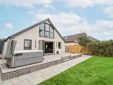 A house with large windows and an open patio door leading to a paved patio with a hot tub and a grass lawn at Ty Blodau near Prestatyn
