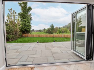 A garden with a stone patio grass lawn and wooden fence viewed through open glass doors at Ty Blodau near Prestatyn