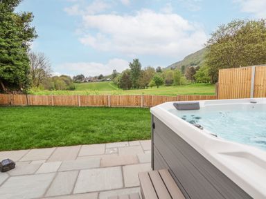 A backyard with a hot tub on a stone patio and a grassy lawn enclosed by a wooden fence near a hill at Ty Blodau near Prestatyn