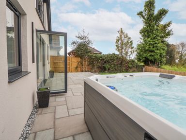 An outdoor patio with a hot tub near a house and a wooden fence with trees in the background at Ty Blodau near Prestatyn