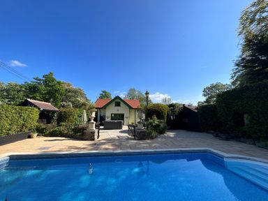 An outdoor swimming pool with a paved patio area in front of a small house surrounded by trees and shrubs at Hafod Lodge in Hensol near Pontyclun