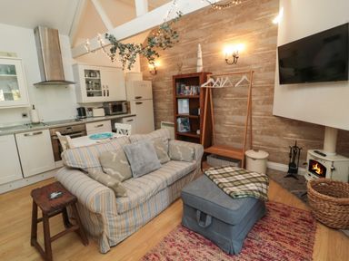 A living room with a striped sofa a wooden stool an ottoman and a kitchen area at The Forge Nr.Seahouses
