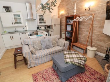 A small living area with a striped sofa, ottoman with a blanket, wooden bookcase, clothing rack, and kitchen in the background at The Forge in Nr Seashouses
