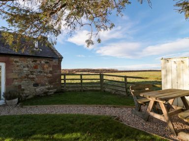 A garden with a wooden picnic table and bench beside a stone building and fencing at The Forge in Nr Seashouses