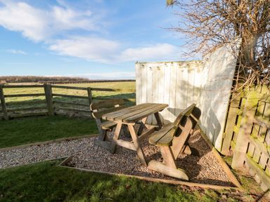 An outdoor seating area with a wooden picnic table and benches on gravel next to a white fence at The Forge Nr Seashouses