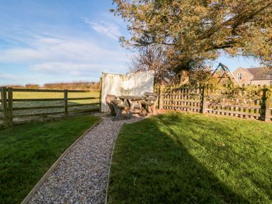 A garden with a gravel path, wooden picnic table and benches, fenced grassy area, and trees at The Forge in Nr Seabouses