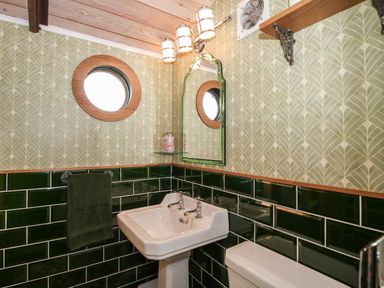 A bathroom with a white sink toilet green tiled walls round window and an arched mirror at GWR Wagon - Victoria Station in Ludlow