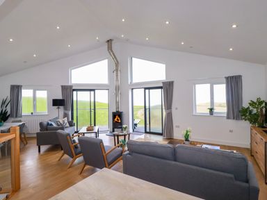 A living room with gray sofas chairs a wood burning stove and large windows at Jacana in Perranporth