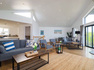 A living room with two blue sofas, two blue armchairs, coffee tables, and a kitchen area at Jacana Perranporth