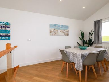 A dining room with a table covered with a tablecloth surrounded by six chairs a plant and beach-themed wall art at Jacana in Perranporth