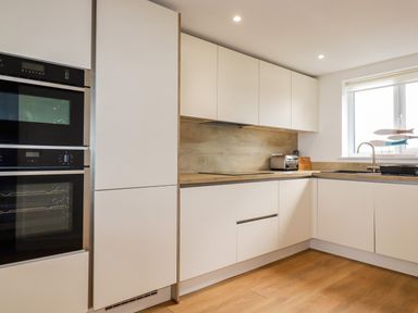 A kitchen with built-in ovens white cabinets a toaster and a sink under a window at Jacana in Perranporth