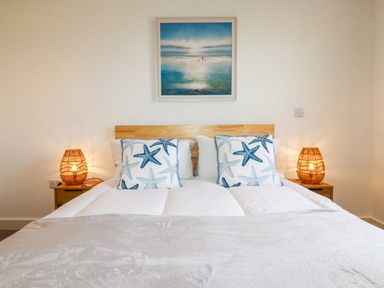 A bedroom with a double bed two starfish pillows wooden bedside tables with lamps and a seascape painting on the wall at Jacana in Perranporth