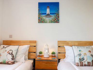 A bedroom with two single wooden beds separated by a wooden nightstand with a lighthouse lamp and small plants and a painting of a lighthouse above the nightstand at Jacana in Perranporth