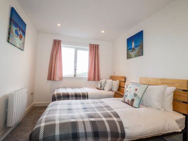 A bedroom with two single beds with plaid blankets and pillows a window with curtains and paintings on the walls at Jacana in Perranporth