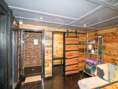 A wooden room with a shower area on the left a sliding wooden door in the center a metal shelf with plants and yoga mats and a cushioned seating area at Orchard Retreat Staycation in Kirton