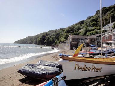 A beach with boats on the sand near a building by the seashore with trees on a hill at Penlee Point in Kingsand