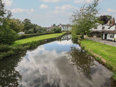 A river with green grassy banks trees and houses in the background at Peony in Eardisland