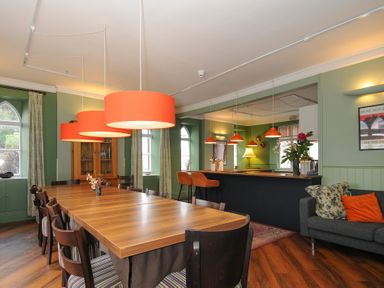 A dining area with a long wooden table and chairs under orange pendant lights next to a bar counter and sofa at Sycamore Apartment in Lifton