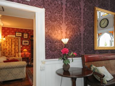 A room with patterned wallpaper a small round table with flowers a wall lamp and a wooden bench with cushions at Sycamore Apartment in Lifton