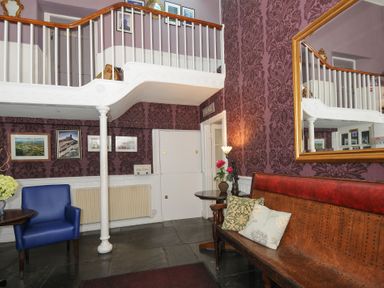 A small seating area with a blue armchair, wooden bench with cushions, side tables, patterned wallpaper, framed pictures, and a balcony railing at Sycamore Apartment in Lifton