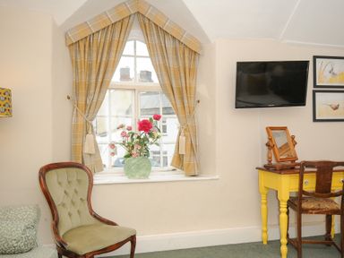 A room with a green upholstered chair yellow desk with a mirror flowers on the window sill and a wall-mounted TV at Sycamore Apartment in Lifton