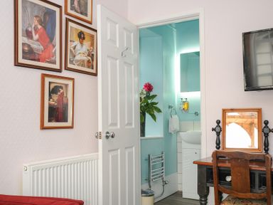 A room with framed paintings on the wall a wooden desk and chair a television and an open door showing a bathroom with a vase of flowers at Sycamore Apartment in Lifton