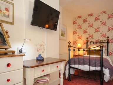 A bedroom with floral wallpaper a metal bed a wooden desk with a blue vase and a wall-mounted television at Sycamore Apartment in Lifton