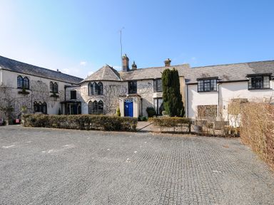 A white building with arched windows and a blue door surrounded by hedges and a paved parking area at Sycamore Apartment in Lifton