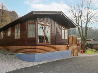 A wooden cabin with multiple windows next to a gravel path and trees at Swifts Reach Lodge Kirkstone 10 Limefitt Park near Windermere
