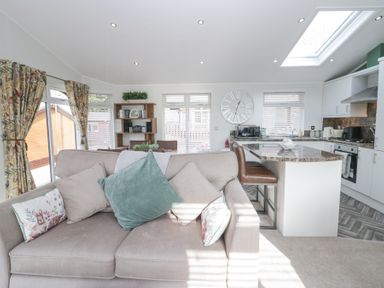 A living room with a sofa and cushions next to a kitchen island with chairs and a wall clock at Swifts Reach Lodge Kirkstone 10 Limefitt Park near Windermere
