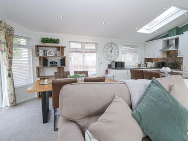 An open plan living room with a beige sofa and green pillow next to a dining table with brown chairs and a kitchen area with white cabinets at Swifts Reach Lodge in Kirkstone 10 Limefitt Park near Windermere