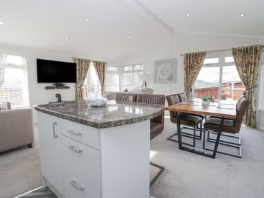 An open living and dining area with a kitchen island in the foreground and a dining table with chairs near windows with patterned curtains at Swifts Reach Lodge in Kirkstone 10 Limefitt Park near Windermere
