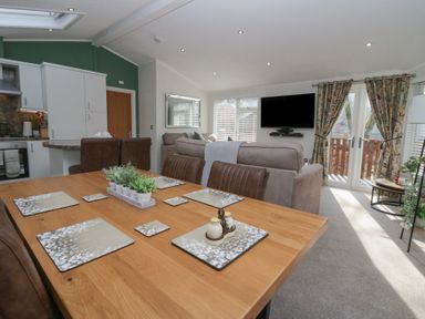 A dining table with place mats and plants adjacent to a living room with sofas a wall mounted tv and glass doors with curtains at Swifts Reach Lodge in Kirkstone 10 Limefitt Park near Windermere