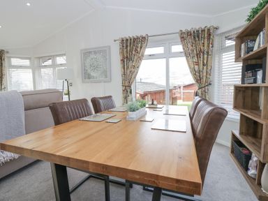A dining area with a wooden table and brown chairs next to windows with floral curtains and a wooden bookshelf at Swifts Reach Lodge Kirkstone 10 Limefitt Park near Windermere