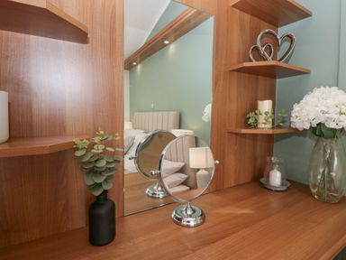 A wooden dressing table with a mirror a small round mirror a black vase with green leaves a glass vase with white flowers a candle and decorative metal hearts at Swifts Reach Lodge Kirkstone 10 Limefitt Park near Windermere