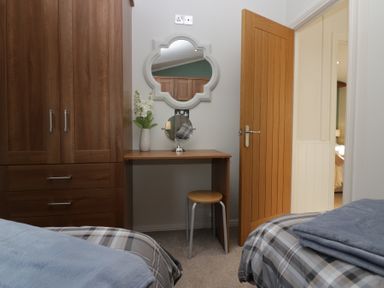 A bedroom with two beds, a wooden wardrobe, a small desk with a round mirror and vase, and an open wooden door at Swifts Reach Lodge Kirkstone 10 Limefitt Park near Windermere