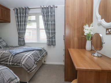 A bedroom with a single bed next to a window with curtains and a wooden dressing table with a mirror and flowers at Swifts Reach Lodge Kirkstone 10 Limefitt Park near Windermere